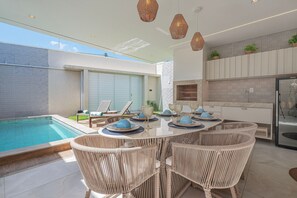 A modern outdoor dining area with wicker furniture, a pool, and a kitchen with a brick backsplash.