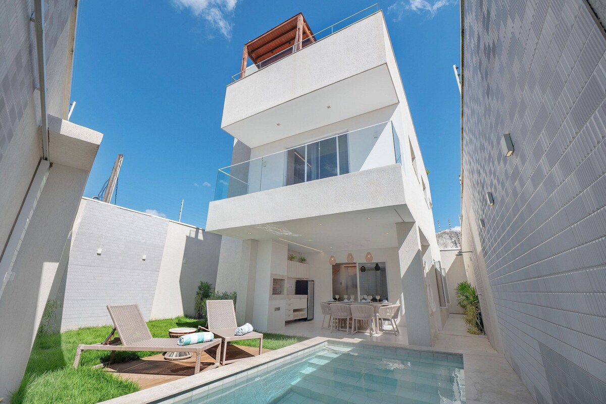 Ein modernes, zweistöckiges Gebäude mit Glasbalkon, Poolbereich mit Liegestühlen und kleiner Terrasse mit Tisch und Stühlen.