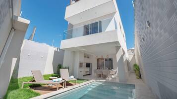 Ein modernes, zweistöckiges Gebäude mit Glasbalkon, Poolbereich mit Liegestühlen und kleiner Terrasse mit Tisch und Stühlen.