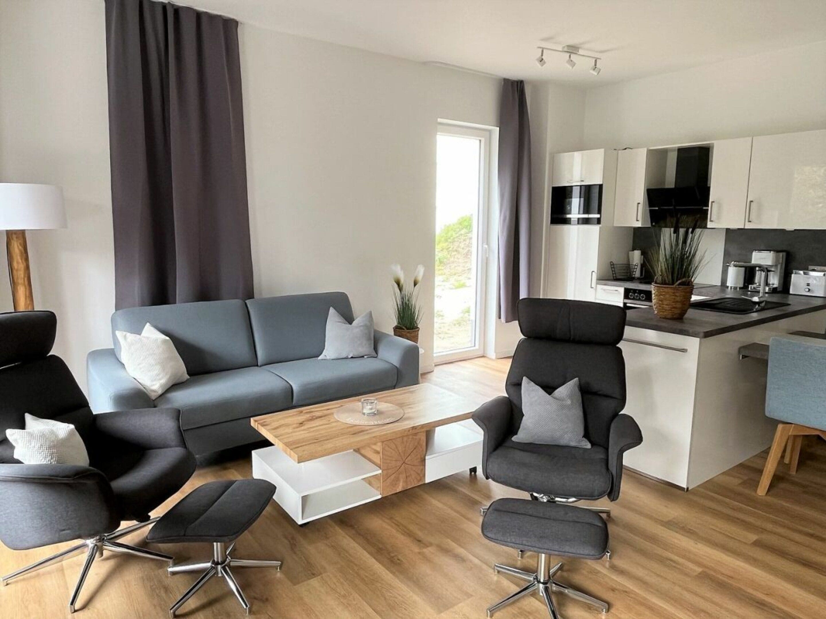 A modern living room with a grey sofa, black armchairs, a wooden coffee table, and a kitchen area with white cabinets and appliances.