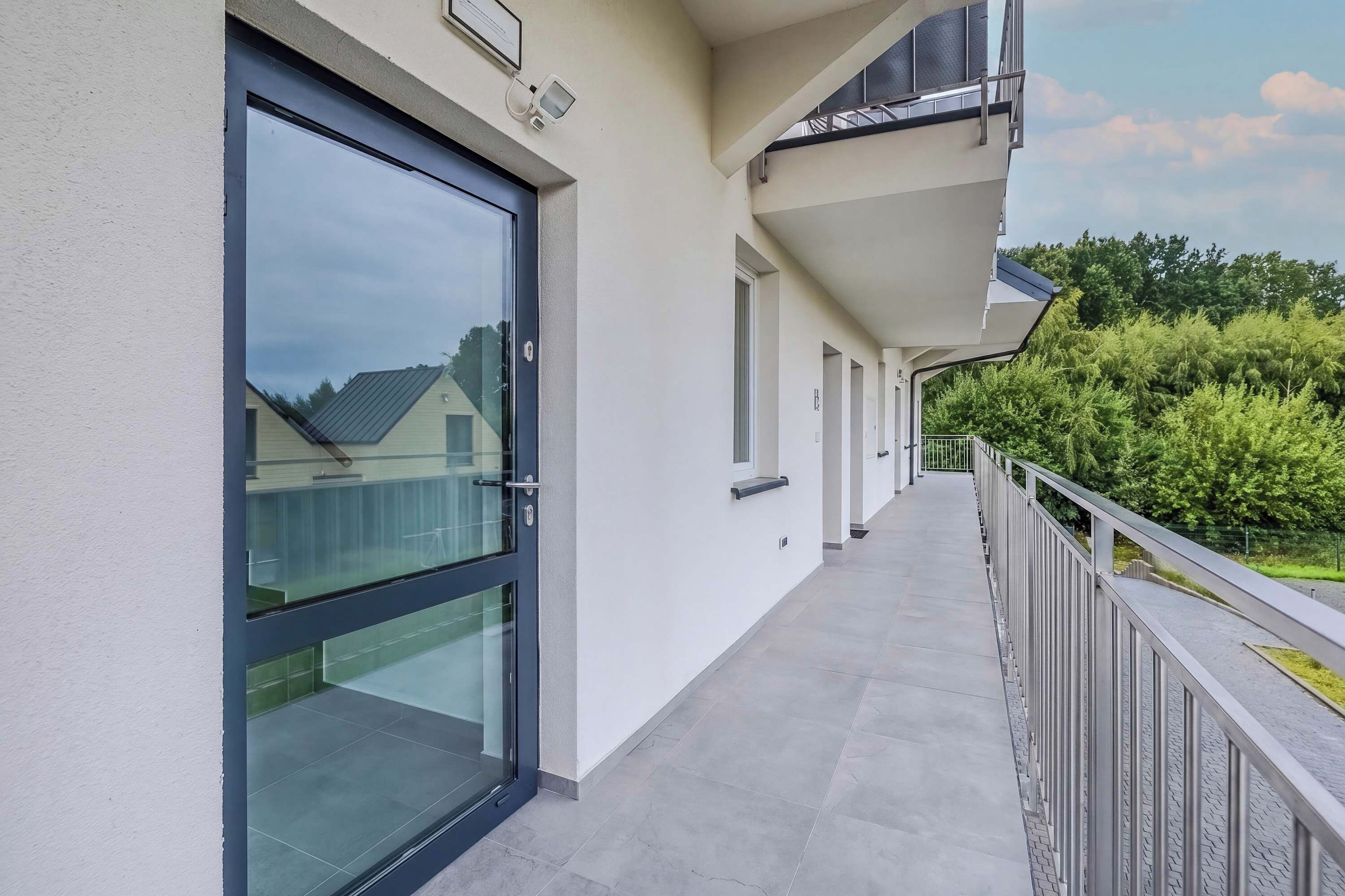 Un pasillo con puerta de cristal que da a un balcón, barandilla metálica y vista a zona residencial con árboles y casas.