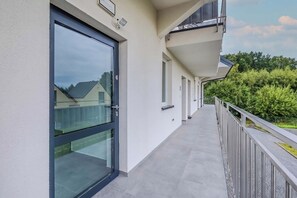A corridor with a glass door leading to a balcony, a metal railing, and a view of a residential area with trees and houses. - Rusinowo | Bosmańska 2 | Cosy Apartment | Balcony | Close to the sea (Rusinowo)