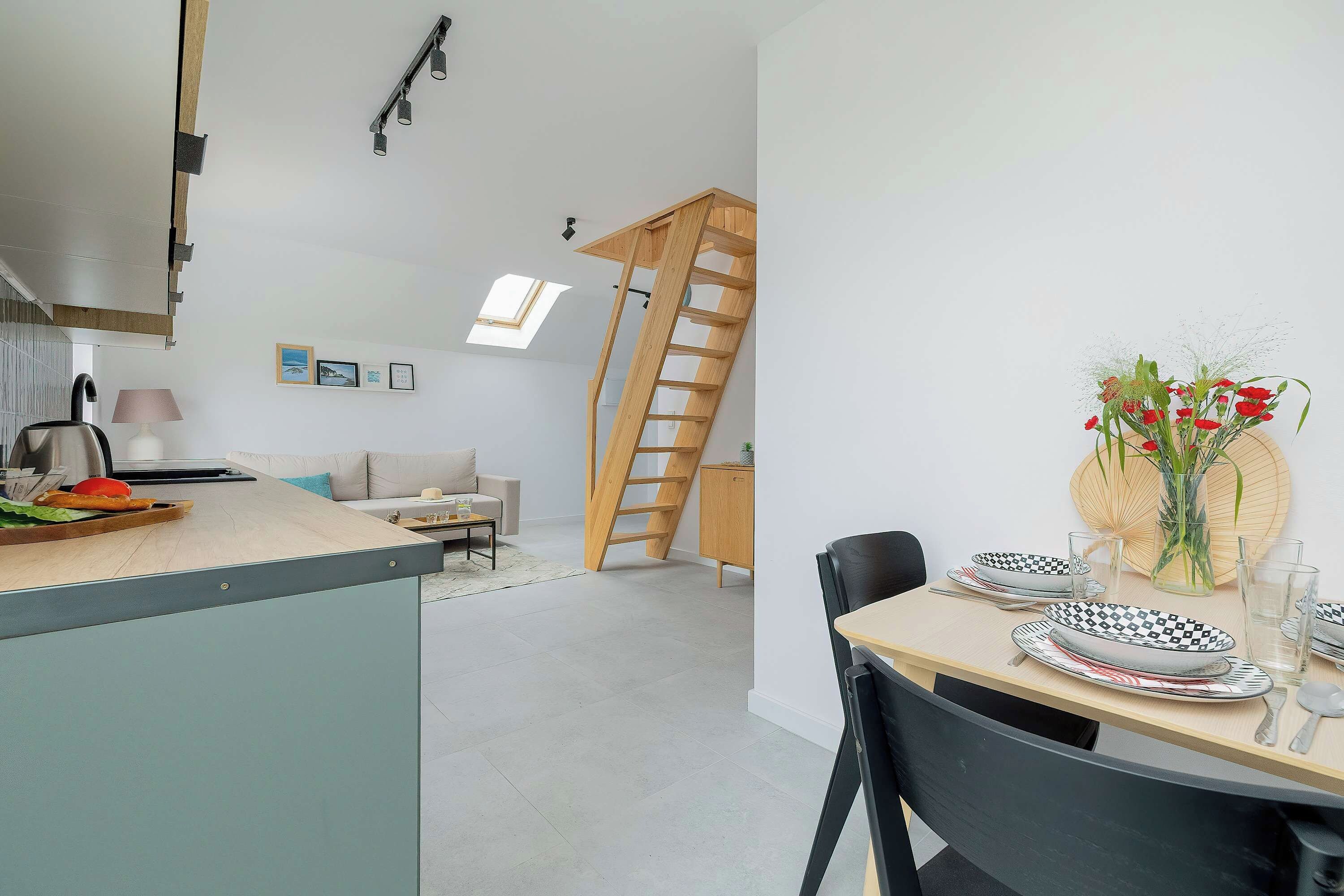 Una cuina moderna amb zona de menjador, un dormitori amb àtic accessible per una escala de fusta, i una taula de dinar coberta amb plats i un ram de flors.