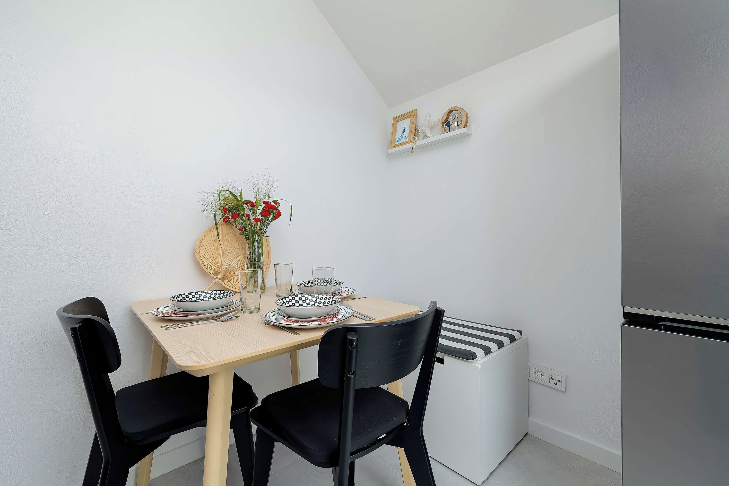 Un comedor con una mesa dispuesta para dos personas, sillas negras, un jarrón con flores rojas y un estante con adornos.