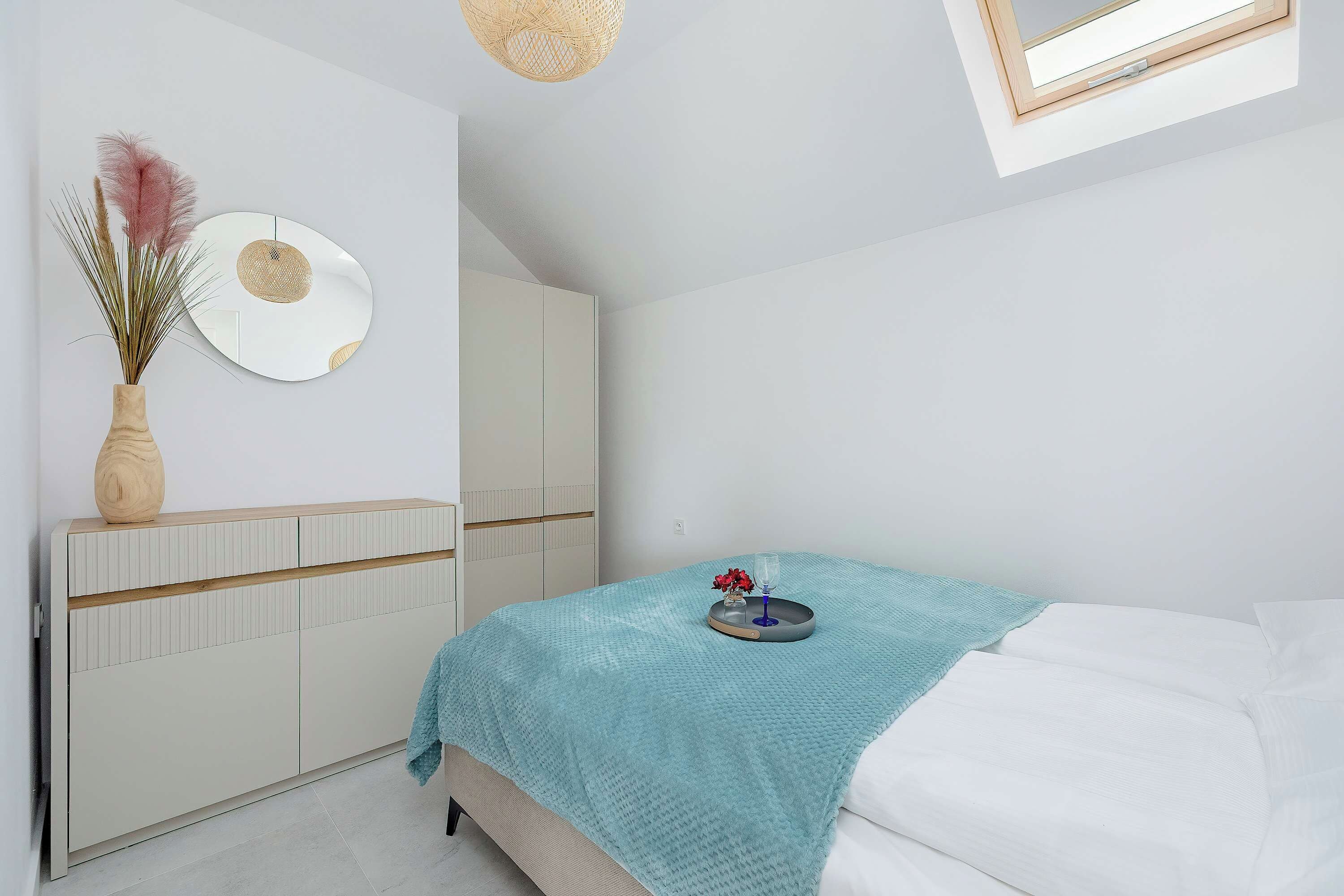 Ein Schlafzimmer mit einem Bett, einem Nachttisch mit einer Vase und einem Glas, einem Kleiderschrank, einem Spiegel und einem Fenster.