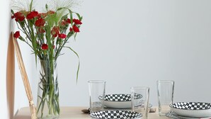 Una mesa de comedor con un jarrón de flores rojas, vasos transparentes y platos con estampado blanco y negro.