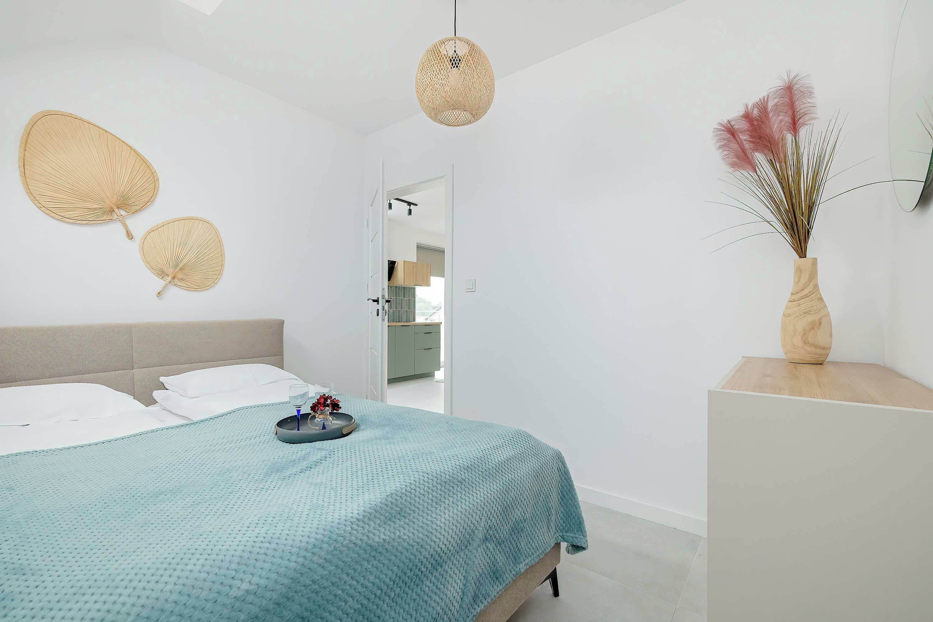 Une chambre à coucher moderne avec un lit, une table de chevet, un vase contenant des plantes séchées et un luminaire suspendu.