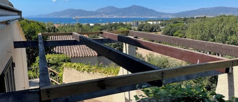 A terrace with a view of the sea and mountains, a wooden railing, and a building with a tiled roof.