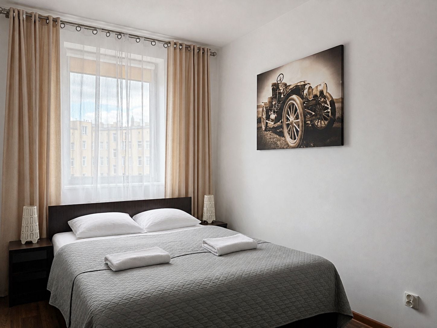 Habitación de hotel con una cama grande, dos mesitas de noche, un ventanal con cortinas y un cuadro colgado en la pared de un coche antiguo.