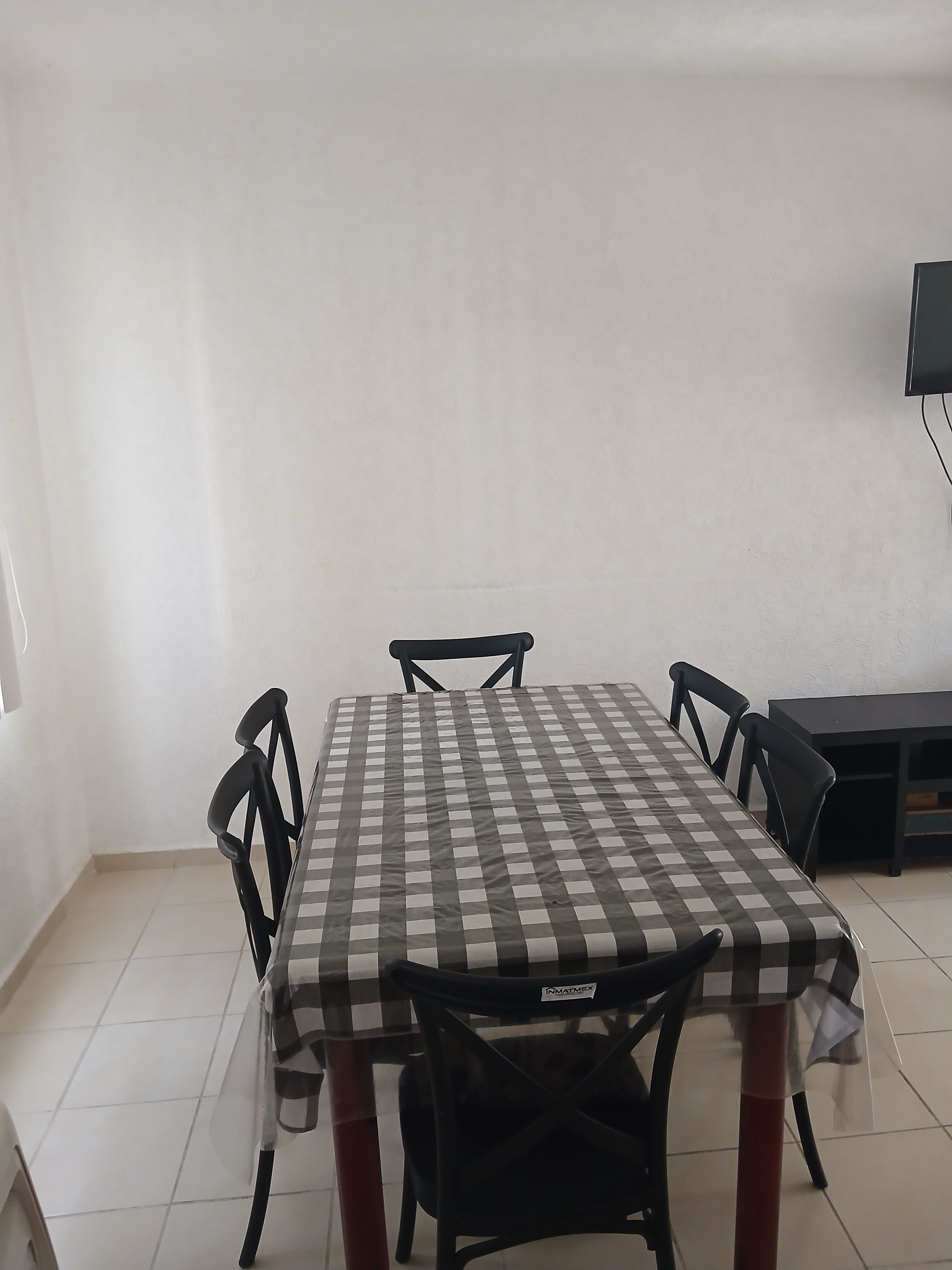 Un comedor con mantel a cuadros, sillas negras y un televisor en la pared.