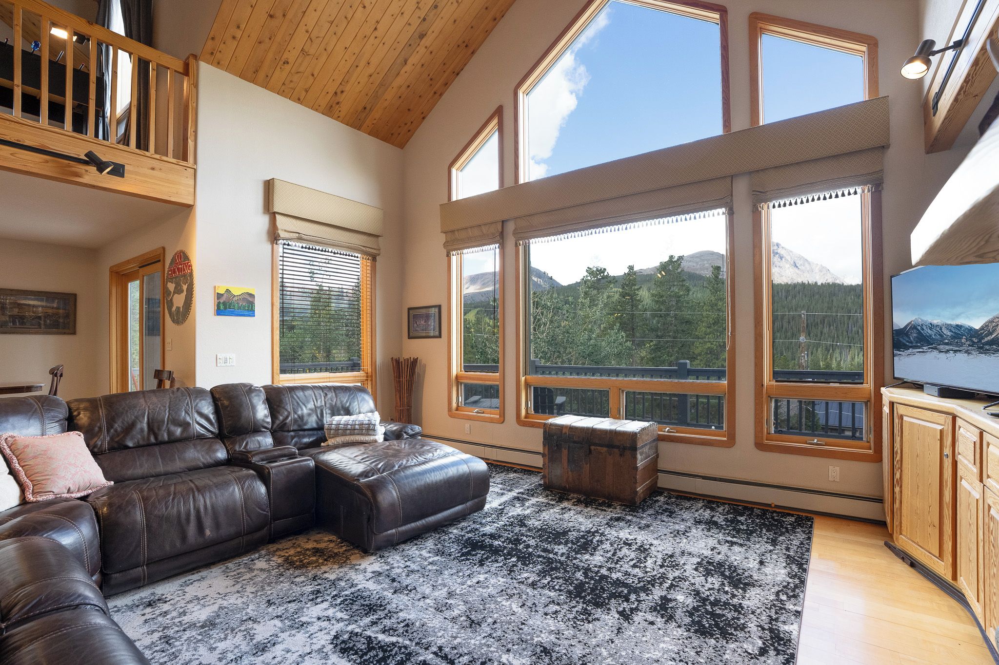 Ein geräumiges Wohnzimmer mit einer ledernen Eckcouch, einem großen Fenster mit Blick auf die Berge und einem Fernseher auf einem Holzschrank.