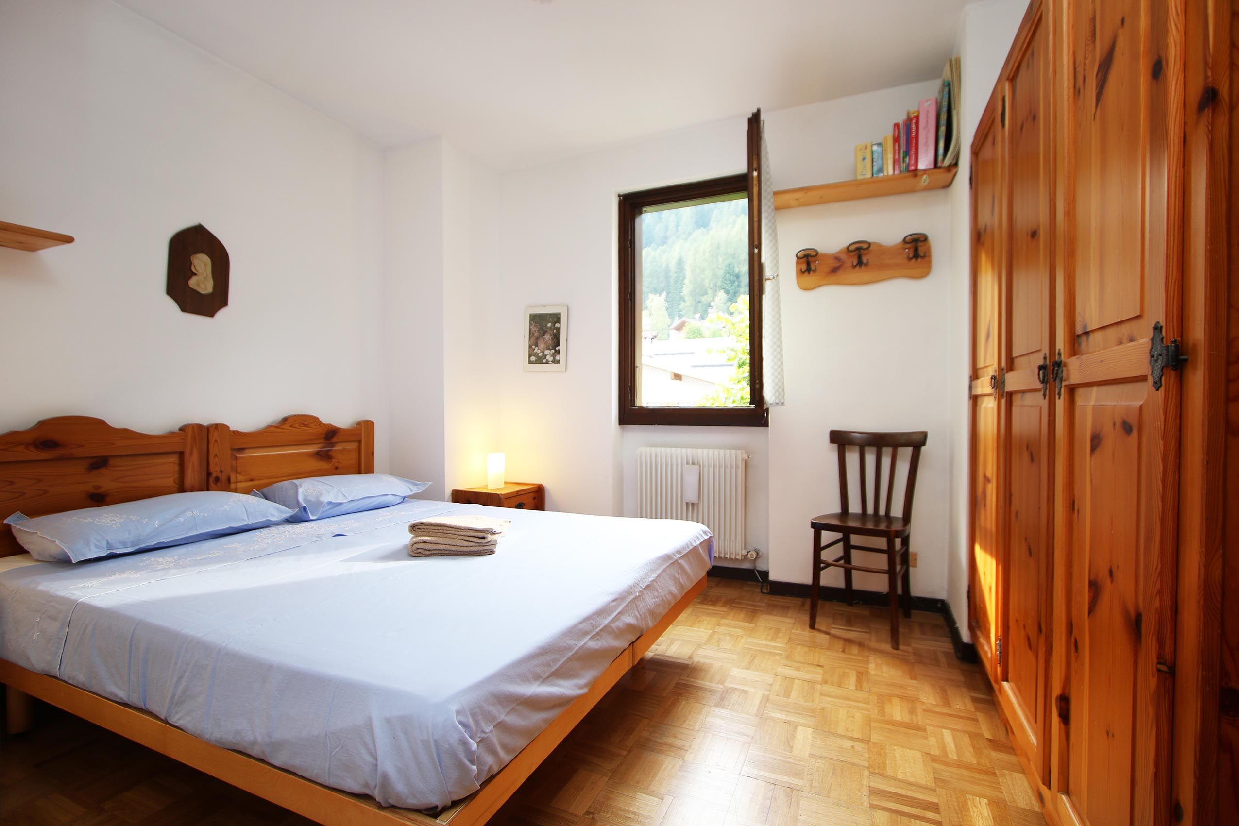Une chambre à coucher avec un lit en bois, une table de chevet avec une lampe, une chaise, un radiateur et une fenêtre donnant sur des arbres.