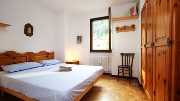 Une chambre à coucher avec un lit en bois, une table de chevet avec une lampe, une chaise, un radiateur et une fenêtre donnant sur des arbres.