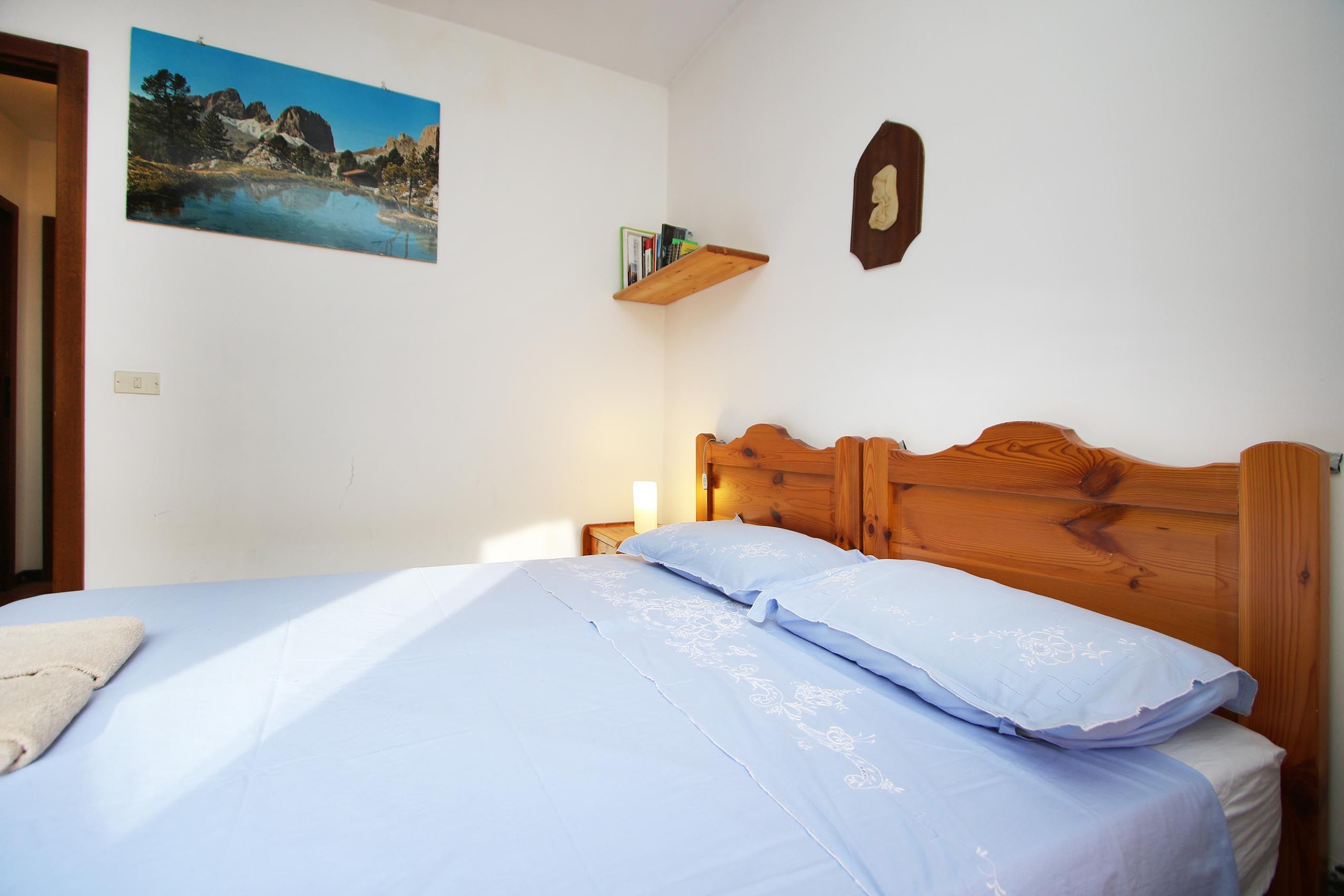 A bedroom with a bed, a nightstand, a wall-mounted shelf with books, and a framed picture.