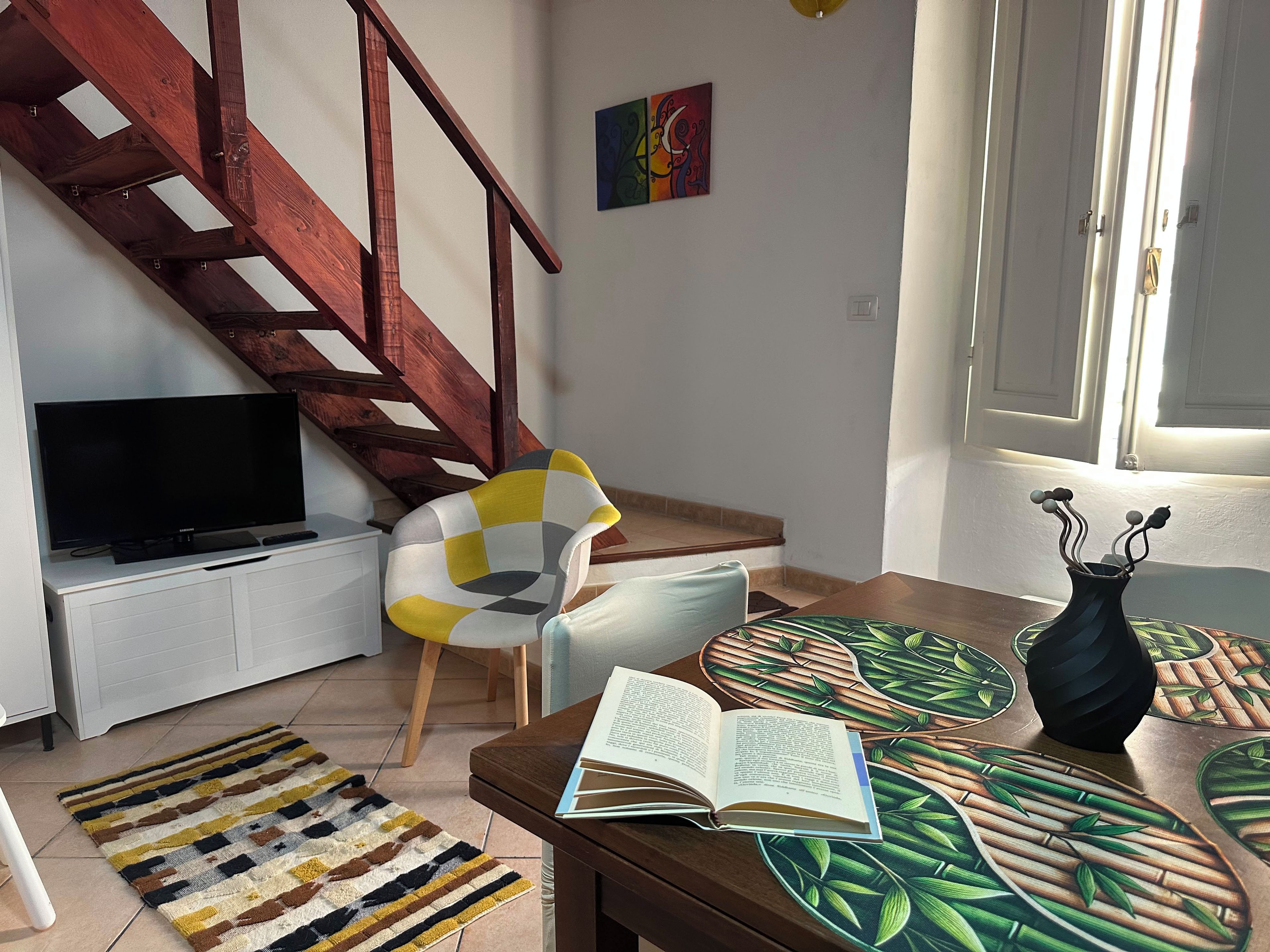 A living room with a TV, a chair, a table with an open book, a patterned rug, and a staircase.