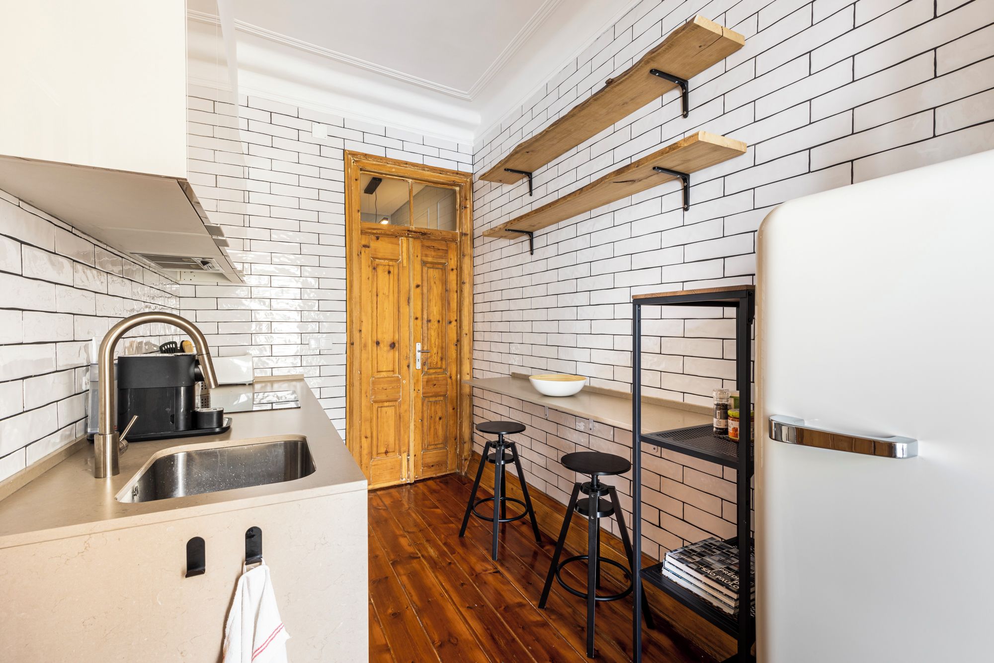 A compact kitchen with a sink, wooden stools, a white refrigerator, and a brick-patterned backsplash.