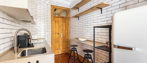 A compact kitchen with a sink, wooden stools, a white refrigerator, and a brick-patterned backsplash.