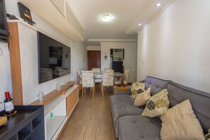 A modern living room with a grey sofa, a TV mounted on the wall, a wooden cabinet, and a dining area with a table and chairs. - Apartment with BBQ 350m from Guilhermina Beach (Praia Grande)
