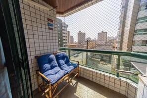 Ein Balkon mit Korbstuhl und Kissen, einer Kachelwand und Blick auf Stadtgebäude durch einen Maschendrahtzaun.