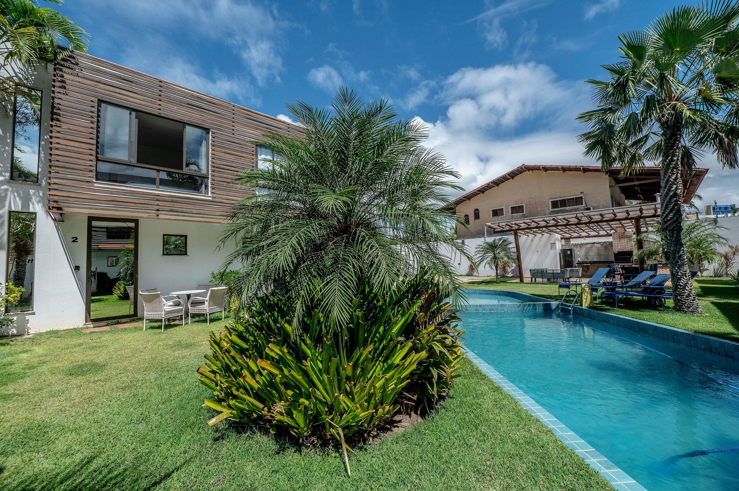 Uma casa moderna de dois andares com piscina, rodeada por vegetação exuberante e palmeiras.