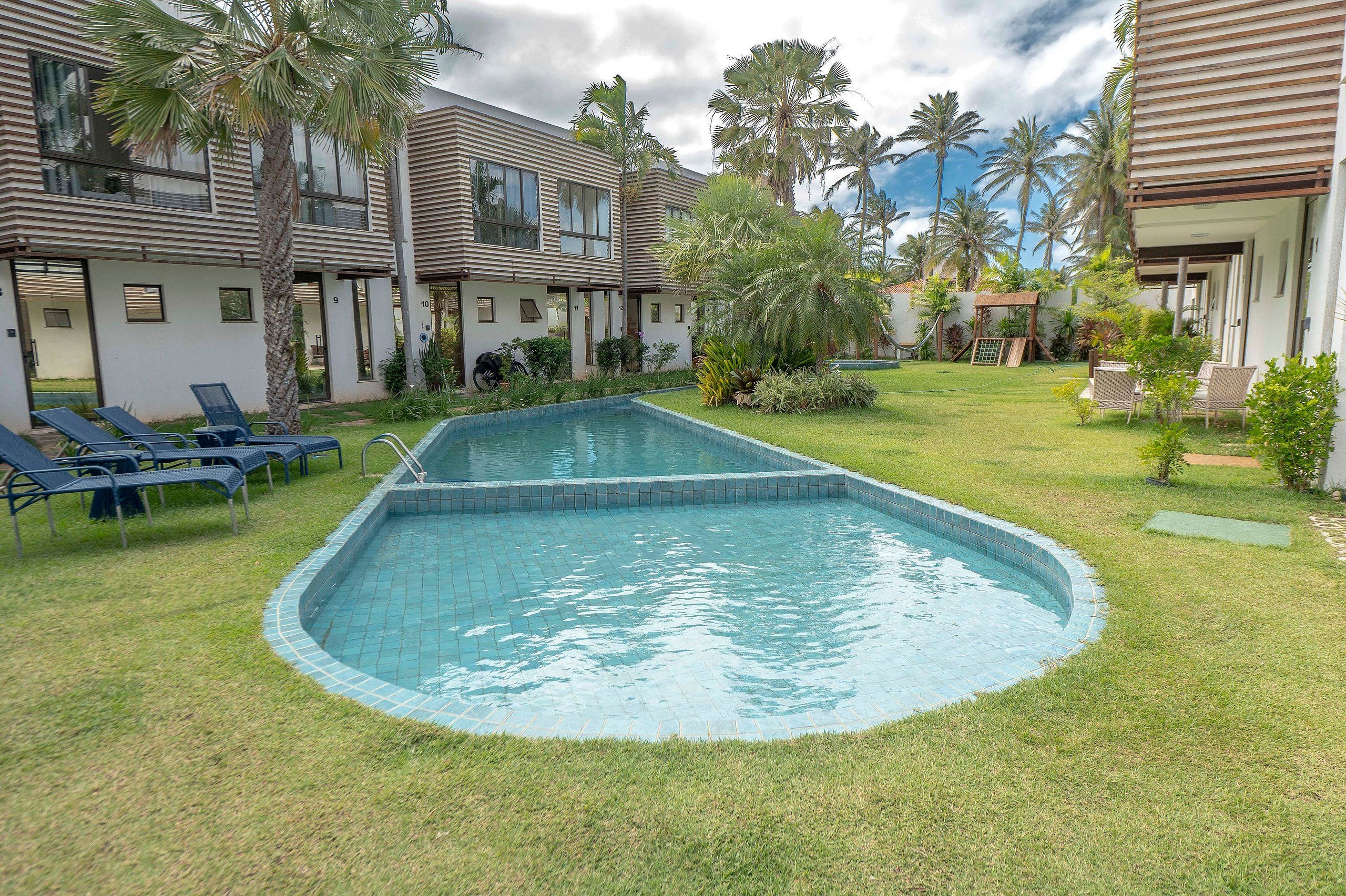 Piscina com borda azulejada, cercada por grama e palmeiras, com um edifício ao fundo.