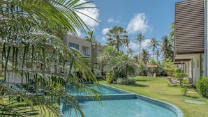 Una piscina con borde de azulejos, rodeada de césped cuidado y diversas plantas tropicales.