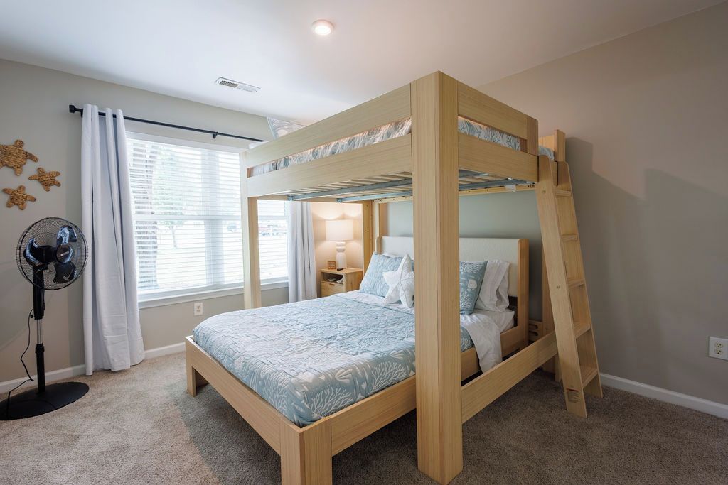 A bunk bed with a ladder, a bedside table, a lamp, and a fan in a room with carpeted flooring and a window with curtains.