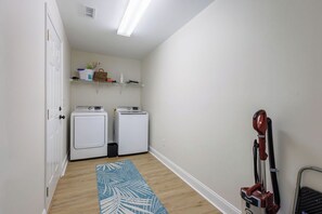 Un cuarto de lavandería con dos lavadoras, una secadora, un aspirador y un estante con diversos objetos.