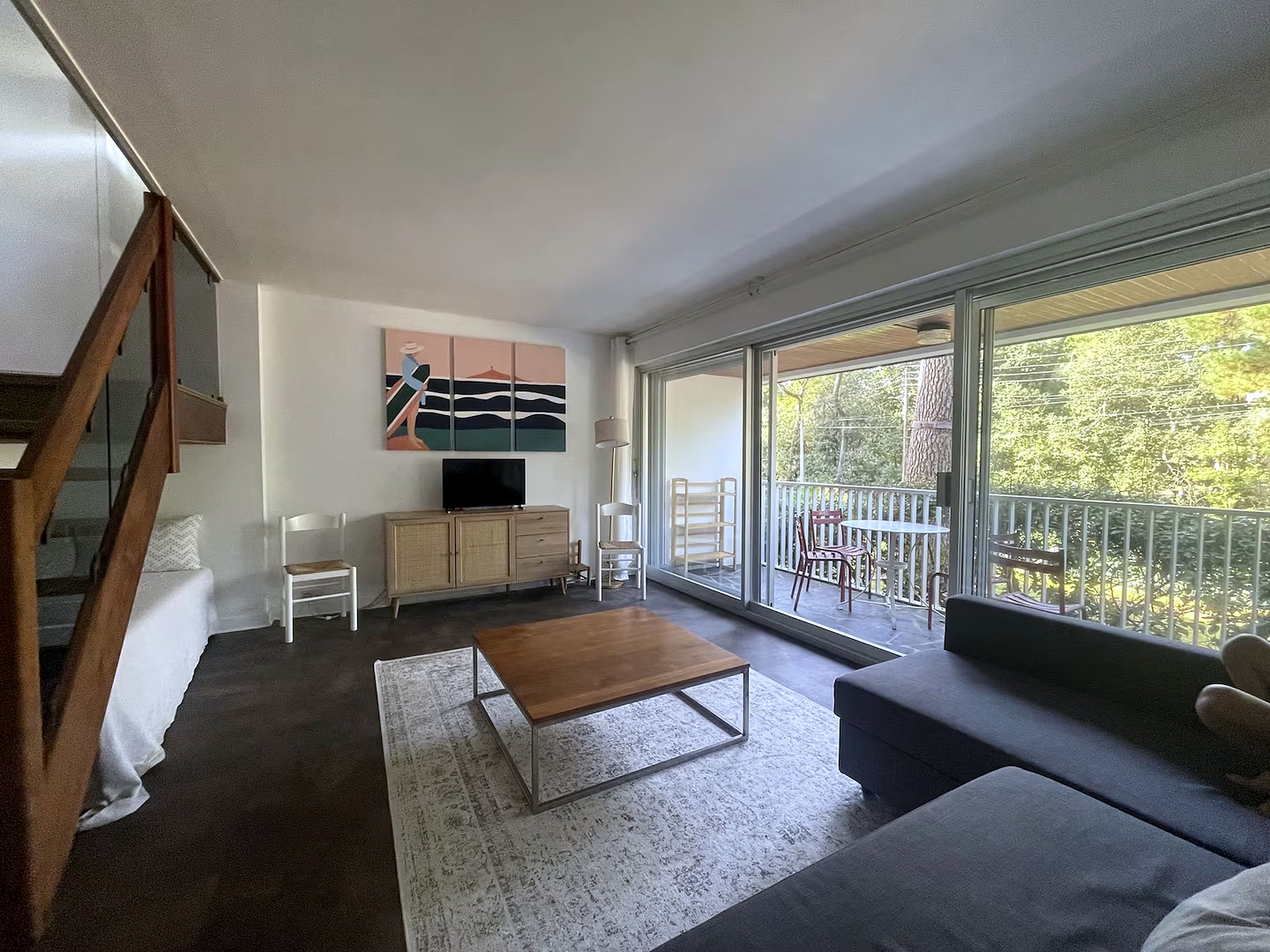 A modern living room with a sofa, a coffee table, a TV on a cabinet, and a balcony with a view of trees.