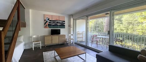 Uma sala de estar moderna com sofá, mesa de centro, TV em um armário e varanda com vista para árvores.