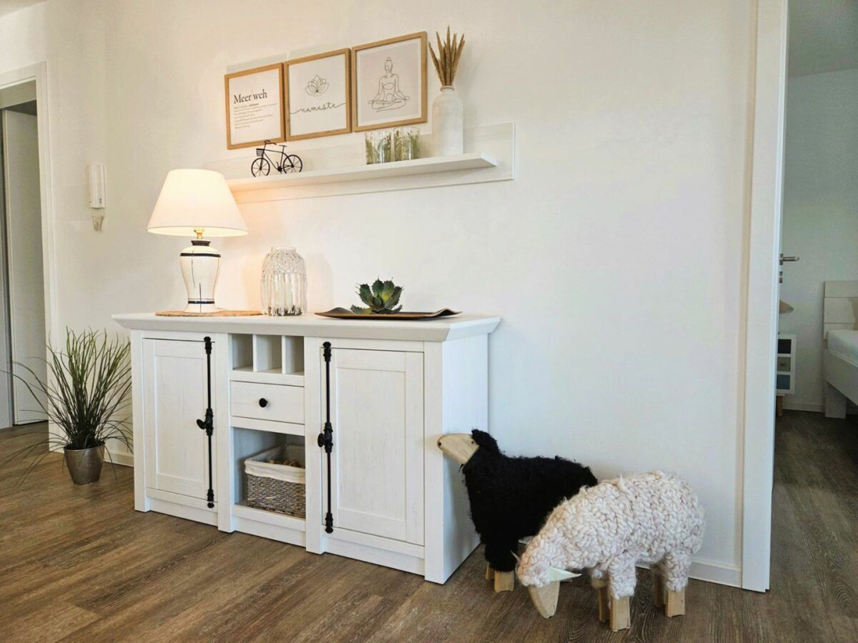 Une console blanche, ornée d’une lampe, d’œuvres d’art encadrées et d’une petite étagère sur laquelle sont disposés des objets décoratifs. Un mouton noir et blanc est posé sur un tabouret en bois, à côté de la console.