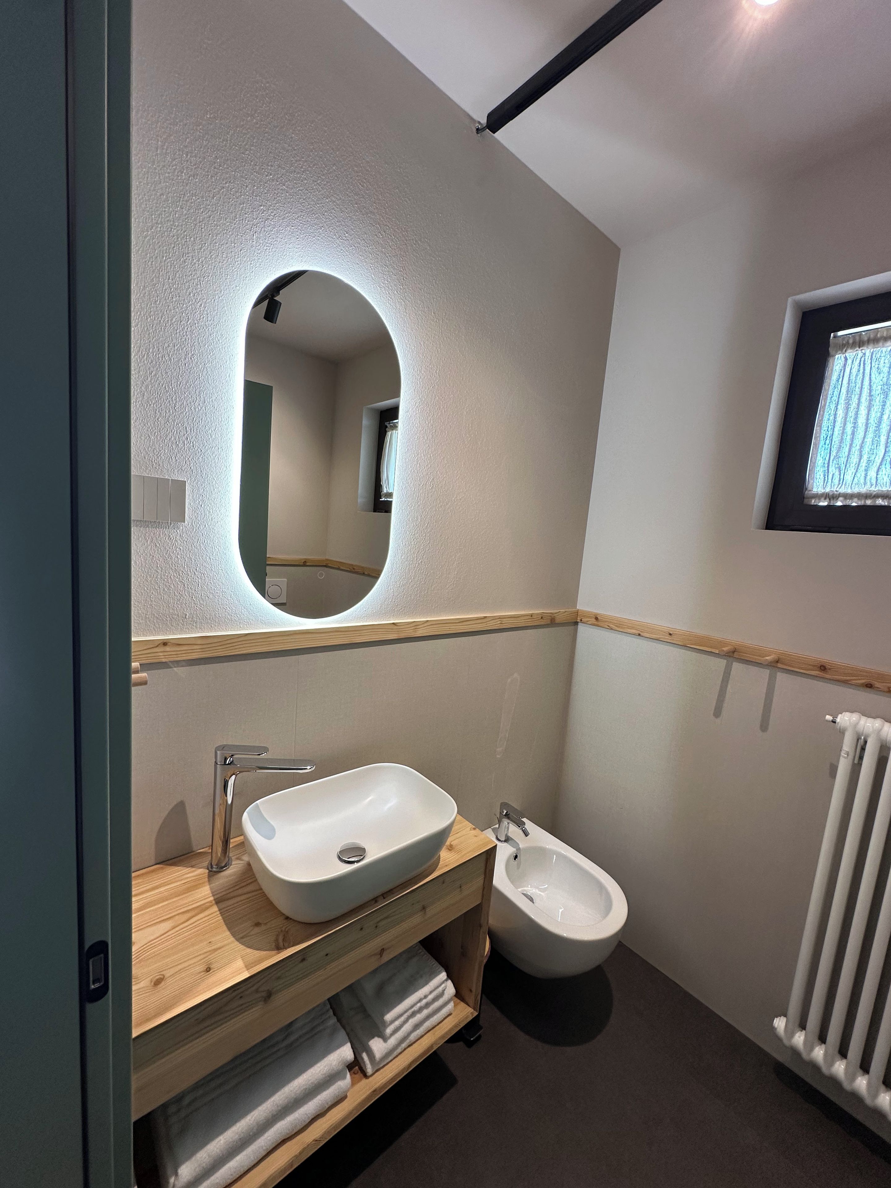 A modern bathroom with a wooden vanity, a large mirror, a bidet, and a radiator.