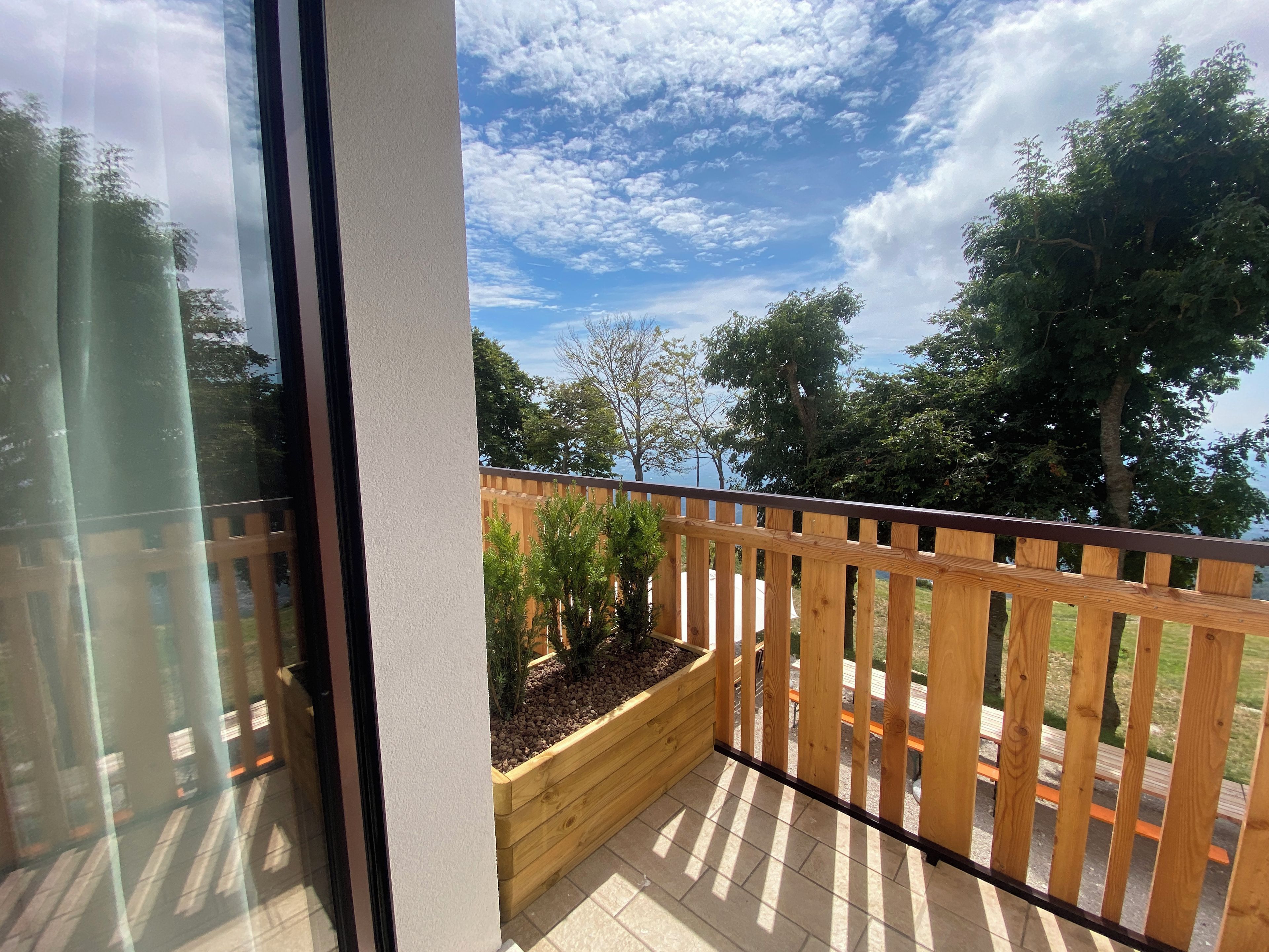 Ein Balkon mit Holzwertkasten, Holzgeländer und Blick auf Bäume und einen klaren Himmel.