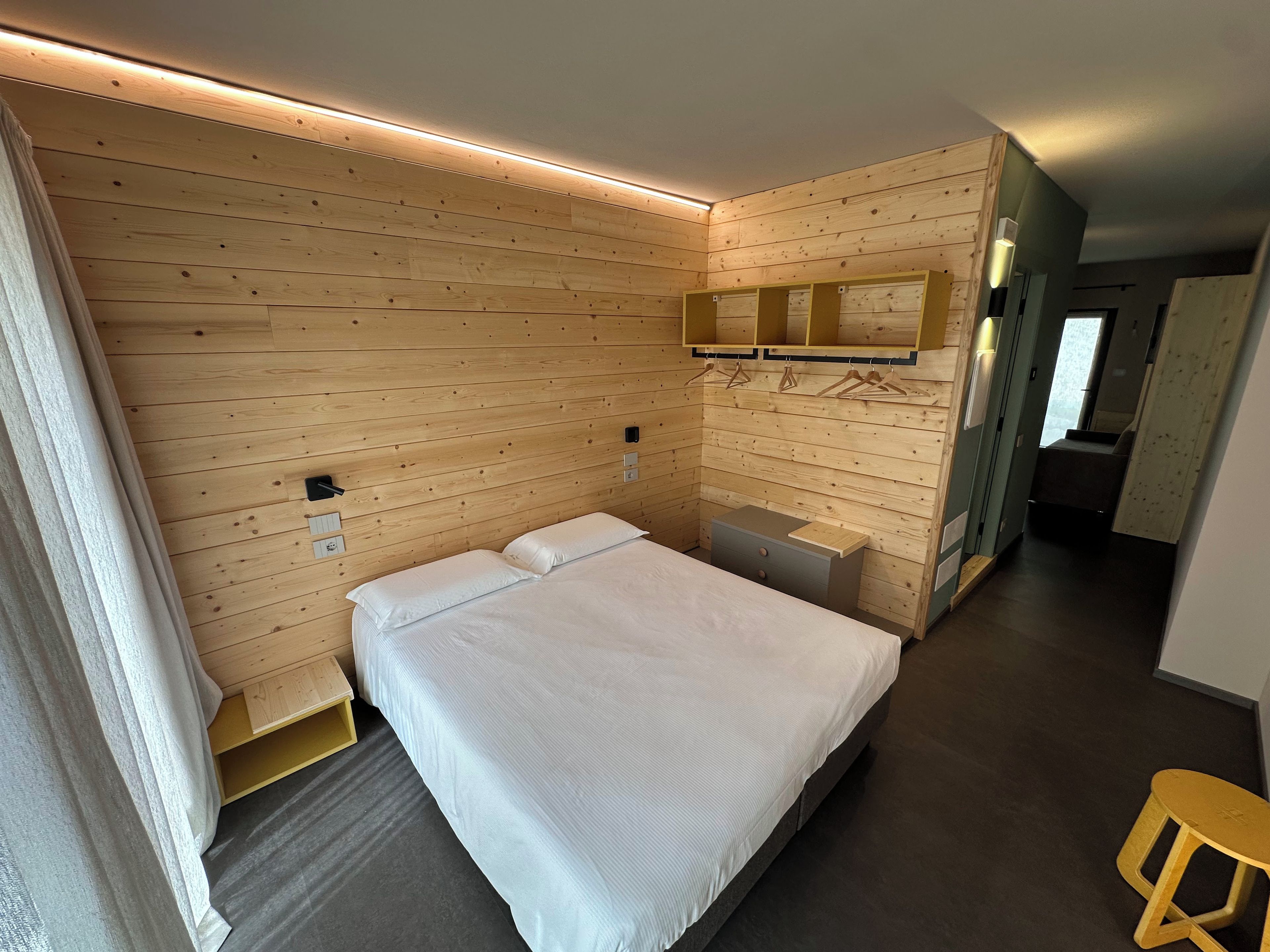 A hotel room with a wooden wall panel, a bed with white bedding, a small bedside table, a mirror, and a stool.