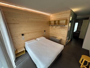 A hotel room with a wooden wall panel, a bed with white bedding, a small bedside table, a mirror, and a stool. - Rifugio San Bernardo (Valdobbiadene)