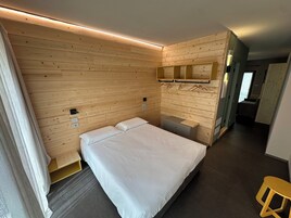 Habitación de hotel con panel de pared de madera, cama con ropa de cama blanca, mesita de noche pequeña, espejo y un taburete.