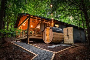 A cabin with a wooden exterior, a large barrel-shaped door, and a deck with string lights. - Sauna + Shower, Spa, Firepit, Gym, BBQ +EV Charger (Blue Ridge)