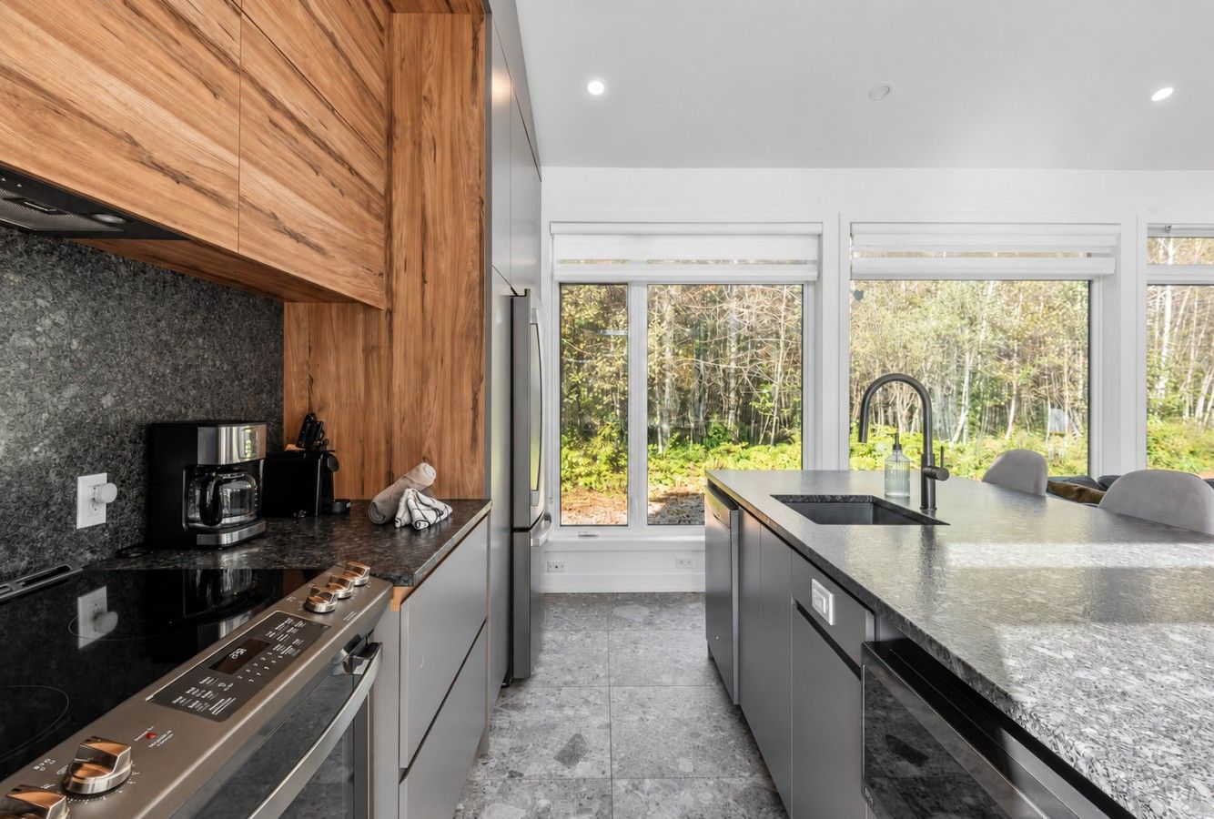 A modern kitchen with a granite countertop, stainless steel appliances, and a view of a forest through large windows.