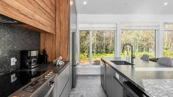 A modern kitchen with a granite countertop, stainless steel appliances, and a view of a forest through large windows.
