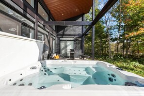 A modern outdoor hot tub area with a wooden ceiling, surrounded by a wooden deck and a glass enclosure, overlooking a forested area.
