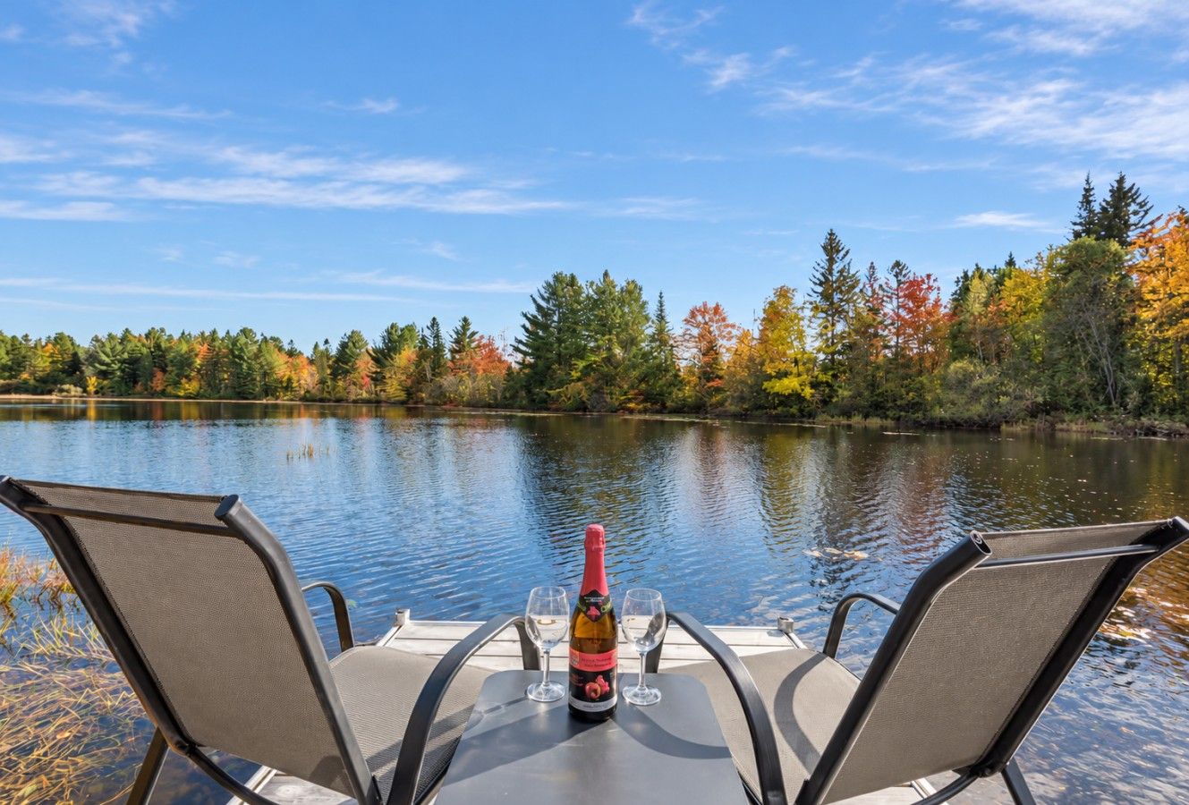 A table with two chairs and a bottle of champagne, overlooking a calm lake with trees in autumn colors.
