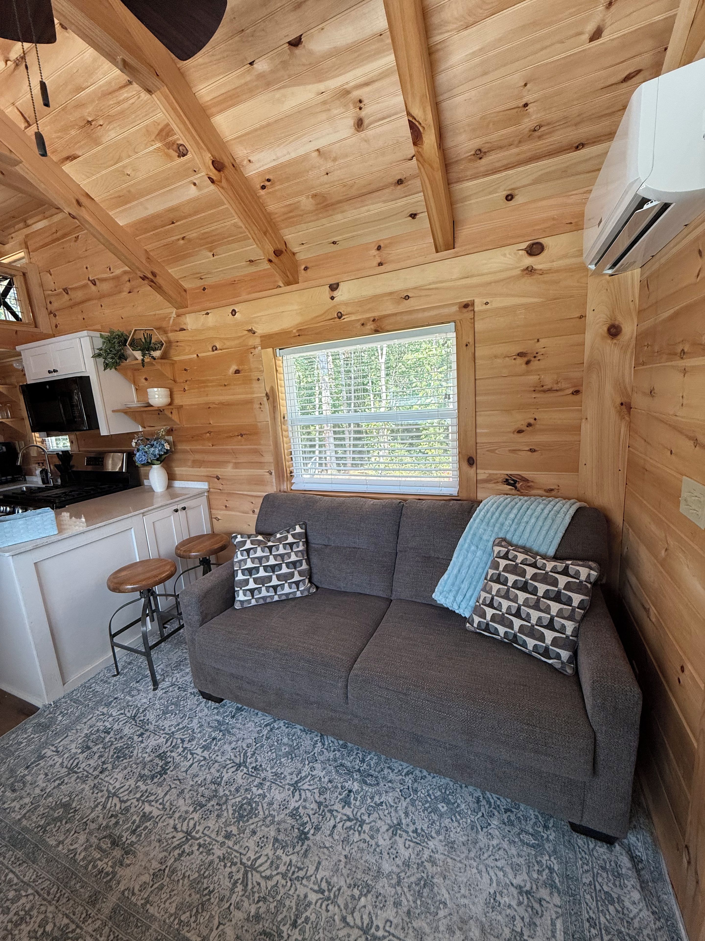 A cozy cabin interior with a sofa, patterned cushions, a kitchen area, and a window with a view.