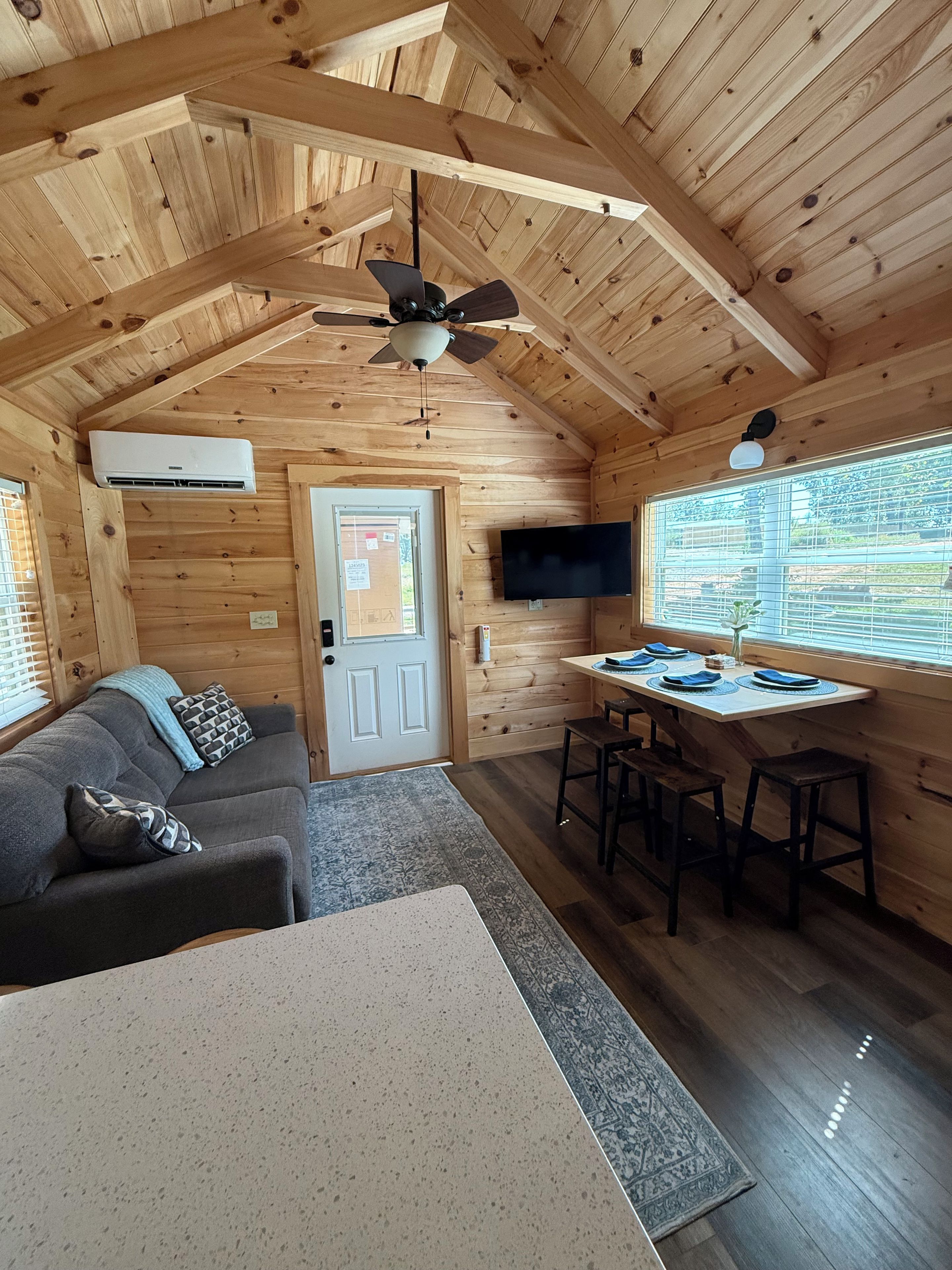 Un intérieur en bois avec un ventilateur de plafond, un téléviseur fixé au mur, un coin repas avec une table et des chaises, et un canapé avec des coussins.