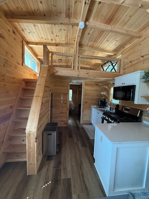 Interior de una cabaña de madera con cocina, comedor y una escalera que sube a un nivel superior.
