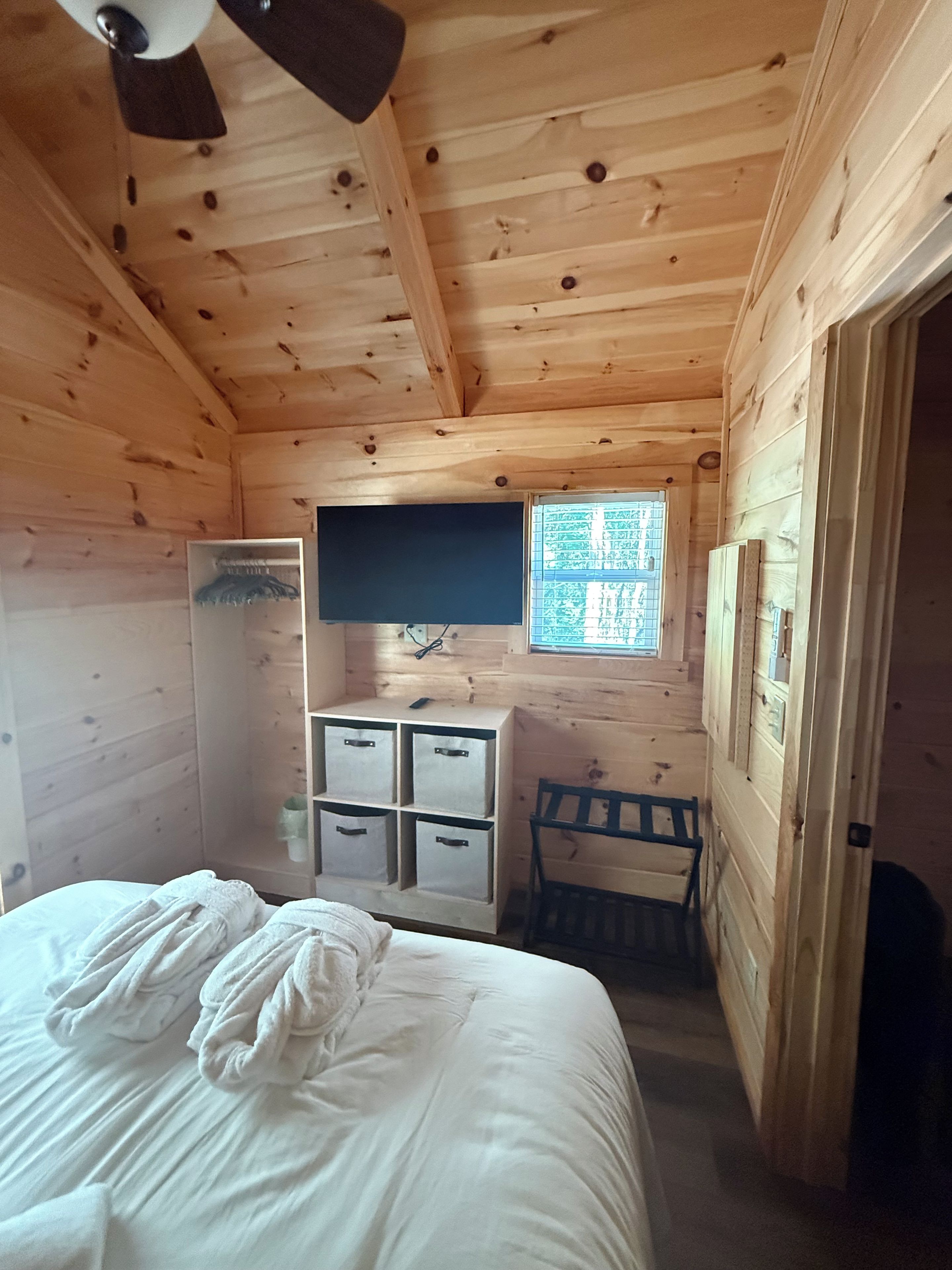A wooden cabin room with a bed, a desk with drawers, a chair, a ceiling fan, and a window.
