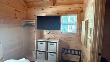 A wooden cabin room with a bed, a desk with drawers, a chair, a ceiling fan, and a window.