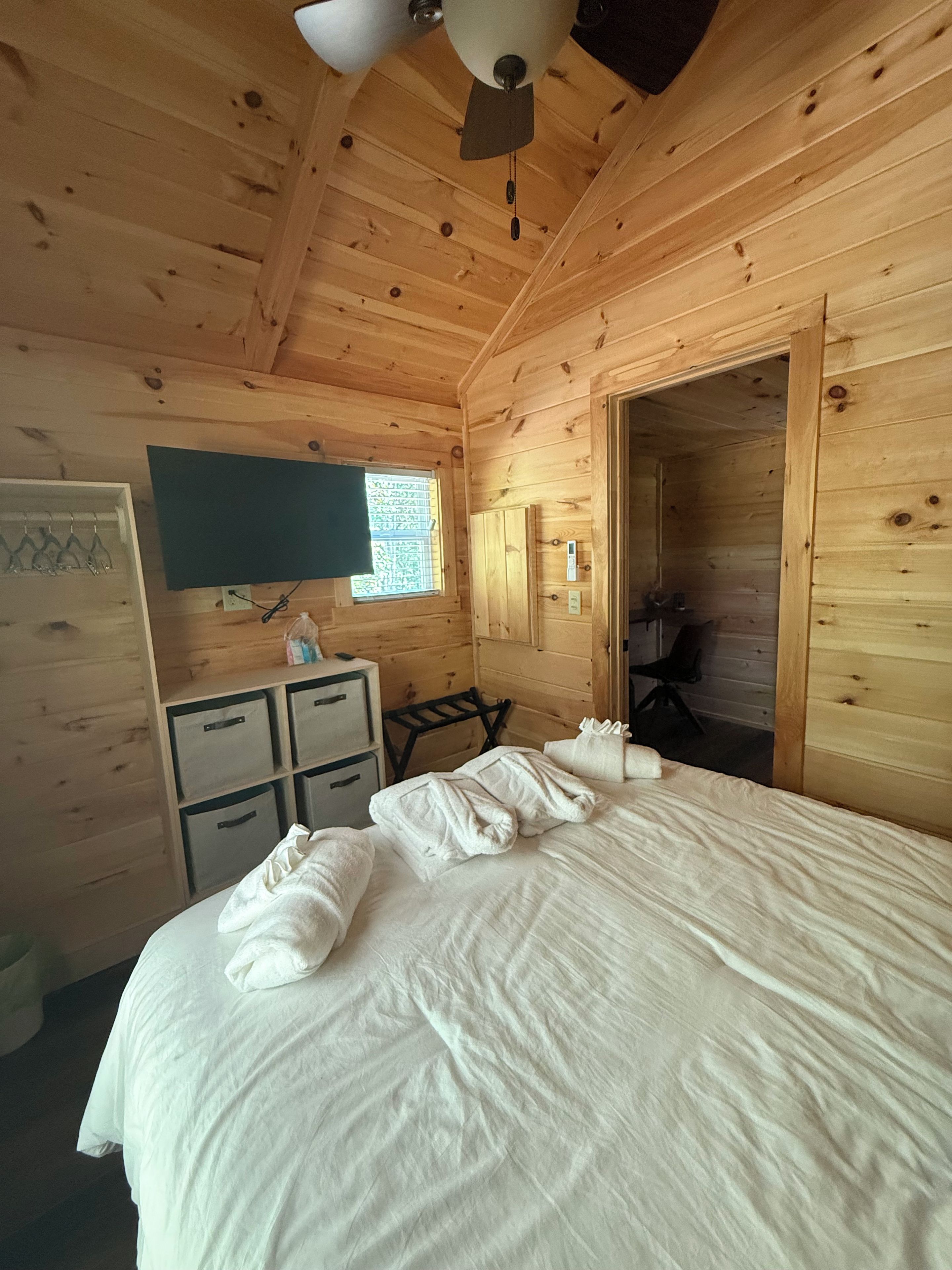 Una habitación tipo cabaña de madera con una cama, un escritorio, una silla, un televisor y una ventana.