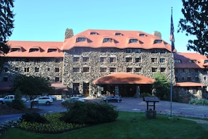 A large stone building with a red roof and multiple windows, surrounded by a well-maintained lawn and parked cars.