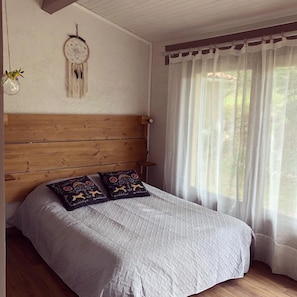 A bed with a wooden headboard, two decorative pillows, a wall-mounted dreamcatcher, and sheer curtains.