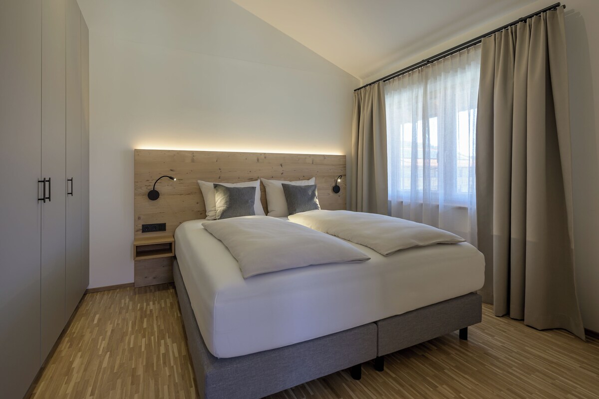 Un dormitorio moderno con cabecera de madera, una cama grande con ropa blanca de cama, dos lámparas de noche y una ventana con cortinas translúcidas.